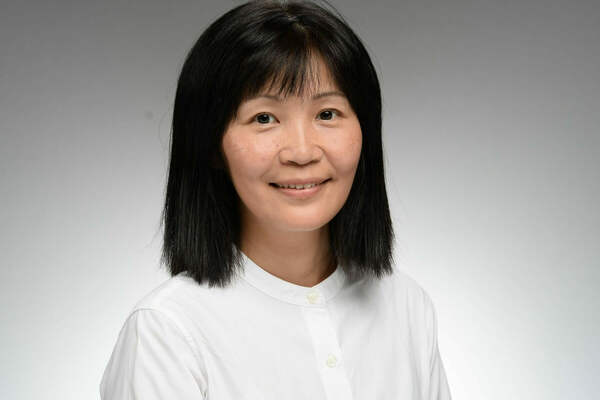 A woman with black, shoulder-length hair and bangs, wearing a white collared shirt, smiles gently at the camera against a grey background.